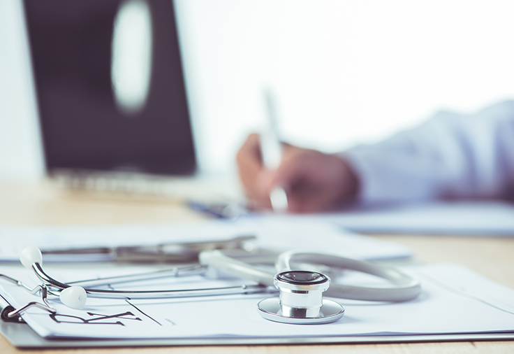 Foreground: A stethoscope on a clipboard. Background: A laptop computer, and a hand writing with a pen.